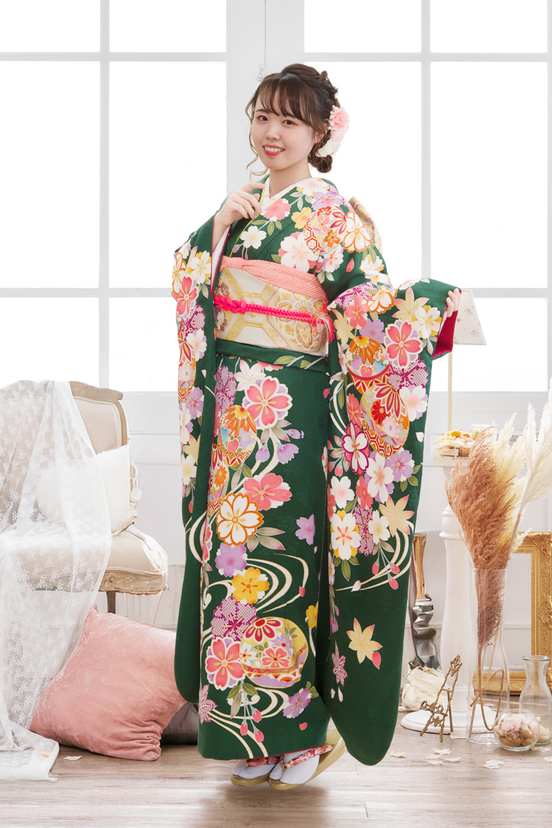 深緑の地にピンクや白の桜が舞い踊る、可憐な古典柄振袖。鞠の紋様と明るい小物使いが二十歳の可愛らしさを引き立てる、清楚で華やかな装い。