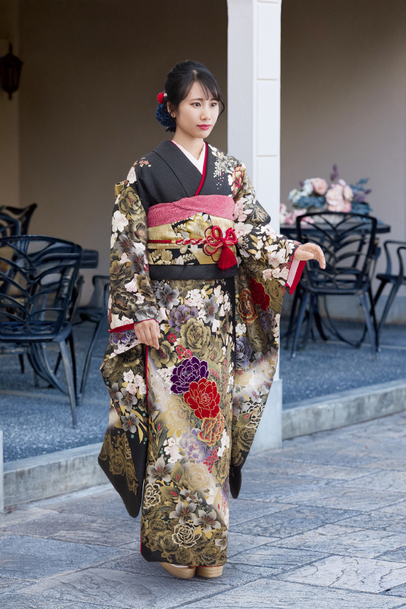重厚な黒地にゴールドの緻密な柄と、深紅や紫の牡丹が浮かび上がるラグジュアリーな振袖。赤の帯締めが全体を引き締める、品格漂う洗練された二十歳の装い。